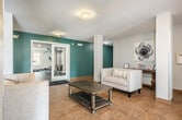 Located at 843 Oak Ave N in Onalaska, Wisconsin, this single-floor community amenity space offers a thoughtfully designed layout that combines a welcoming entrance, comfortable lounge areas, and a dedicated fitness center. The entrance greets visitors through glass-paneled doors framed with rich wood accents, opening into a bright and airy lobby. The lobby features warm tile flooring throughout, neutral-toned walls, and flush-mounted ceiling lighting that creates a clean, inviting atmosphere. A tastefully arranged seating vignette with a tufted sofa, accent chair, and wooden console table adorned with decorative art anchors this welcoming area. The space flows into a spacious open lounge distinguished by bold teal accent walls, additional seating with neutral upholstered sofas, and a central coffee table. Built-in shelving along one wall provides functional storage and display space, while windows allow natural light to filter in and offer views of the surrounding exterior. Accessible through glass doors from the lounge, the fitness center is well-equipped with a treadmill, multi-station cable machine, free weights, and a wall of mirrors that enhance the sense of space. Dark rubber flooring and recessed lighting give the fitness area a practical, professional feel. Together, these areas provide a functional and comfortable community environment.