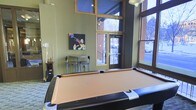 This single-floor recreation space is designed for leisure and social engagement, centered around a full-size pool table that anchors the room as its defining feature. The space boasts impressively high ceilings with an upper band of clerestory windows, allowing abundant natural light to pour in and creating an open, airy atmosphere. Large floor-to-ceiling windows and glass-paneled entry doors line one wall, offering views of the surrounding outdoor environment and further enhancing the sense of openness. The walls are finished in a warm olive-green tone, lending the space a relaxed, inviting character. A dedicated cue rack keeps pool accessories neatly organized and within reach. A small bar-height table with stools provides a comfortable spot for spectators or casual conversation. Colorful pop-art wall décor, including a bold trumpet musician painting, adds a creative and energetic touch to the room's personality. The combination of practical amenities, thoughtful design, and plentiful natural light makes this recreation room a welcoming gathering space suited for both competitive play and casual socializing.