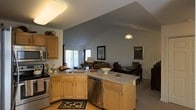 This single-story space showcases an open-concept floor plan that seamlessly connects the kitchen, dining, and living areas into one cohesive, flowing environment. The standout architectural feature is the dramatically vaulted ceiling, which rises to a sharp peak and lends a strong sense of height and openness throughout the main living space.

The kitchen is well-appointed with light wood cabinetry, granite countertops, and stainless steel appliances, including a range and dishwasher. An L-shaped layout with an extended island or peninsula provides generous counter space and creates a natural boundary between the kitchen and the adjoining areas. A double sink is integrated into the main counter run, and a decorative rug anchors the kitchen floor zone.

The dining area sits adjacent to the kitchen and features a formal table set with chairs, accompanied by framed artwork on the walls. The living area beyond is furnished with comfortable seating and benefits from natural light streaming in through a sliding glass door, which also offers a view to the outdoors. Neutral gray-blue wall tones and light-toned flooring create a calm, cohesive ambiance throughout the entire space.
