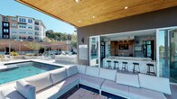 This expansive outdoor amenity space centers on a resort-style pool deck designed to deliver a high-end recreational experience. The pool itself features a striking infinity edge that visually merges with the rolling, tree-covered hillside beyond, creating a seamless connection between the built environment and nature. Integrated water jets and in-pool sun loungers add to the resort ambiance, while rows of sleek chaise lounges line the surrounding deck for sunbathing.

A generously sized covered pavilion anchors one end of the pool deck, supported by slender columns and finished with a warm wood-plank ceiling that adds a natural, refined aesthetic. Beneath the pavilion, large-scale sectional sofas in neutral tones provide a comfortable lounge area that flows effortlessly between shade and sun. Folding glass doors open directly from the pavilion into an interior bar and lounge space, which features a marble-topped bar counter with contemporary black barstools, wood-accent wall cladding, and recessed lighting — ideal for social gatherings.

Ornamental desert plantings, including a prominent yucca, punctuate the pool surround, adding sculptural greenery to the hardscape. The entire area benefits from abundant natural light and panoramic views of the surrounding hills, creating an elevated outdoor retreat atmosphere throughout the day.