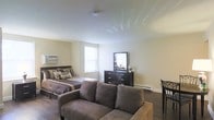 This single-floor studio is thoughtfully arranged to maximize functionality within an open-concept layout that seamlessly integrates the living, sleeping, and dining areas. The main living area features a plush, earth-toned sofa that anchors the space, providing a comfortable lounge area that flows naturally into the sleeping zone. The sleeping area is furnished with a full-sized bed dressed in coordinating neutral linens, complemented by a dark-finished dresser with a mirror that adds depth and practicality to the room. Natural light filters in through a window positioned near the bed, brightening the warm, neutral-toned walls and contributing to an airy, welcoming ambiance. Dark hardwood-style flooring runs throughout, offering a grounding contrast to the light walls and ceiling. A dining area with chairs is tucked to the side, rounding out the multi-functional nature of this efficient layout. The clean, smooth ceiling and baseboard heating are practical features that add to the overall comfort of the space. The understated, neutral color palette and cohesive furnishings create a calm and livable environment well-suited for everyday use.
