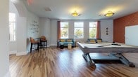 This single-floor social lounge is thoughtfully designed to serve as a dynamic gathering space with a cohesive mid-century modern aesthetic. The open layout is anchored by rich hardwood-style flooring that flows seamlessly throughout, tying together the various activity zones within the room.

The main lounge area features a full-sized billiards table as a central focal point, surrounded by comfortable teal sofas accented with mustard yellow throw pillows. Clusters of bistro-height tables and curved leather chairs provide additional casual seating throughout the space. Large windows dressed with rust-orange drape panels frame views of palm trees and the surrounding outdoor environment, allowing generous natural light to fill the interior.

A wet bar area along one wall is outfitted with dark cabinetry, granite countertops, a farmhouse-style sink, and mosaic tile backsplash. An eye-catching orange geometric-patterned accent wall adds a bold, energetic contrast to the neutral tones of the surrounding walls.

Decorative starburst wall accents, pendant lighting, and thoughtfully curated furnishings contribute to the space's warm, inviting ambiance, making it well-suited for social events and recreational use.