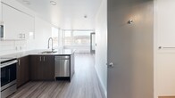 This efficiently designed one-bedroom, one-bathroom unit in Seattle, WA offers a thoughtfully organized single-floor layout with contemporary finishes throughout. The open-concept kitchen and living area serves as the heart of the unit, featuring white upper cabinetry, a peninsula with a gray quartz-style countertop, stainless steel appliances including a refrigerator, range, microwave, and dishwasher, and a classic subway tile backsplash. Light wood-style luxury vinyl plank flooring runs throughout the main living area, complementing the clean, modern aesthetic. Large windows in the living area frame notable outdoor views, including what appears to be a waterway and surrounding hills, flooding the space with natural light.

The bedroom is a comfortable, carpeted room with crisp white walls and a dedicated reach-in closet equipped with a hanging rod, offering practical storage. The bathroom features a full soaking tub and shower combination with chrome fixtures, wood-look vinyl flooring, tile accent walls, and an integrated stacked washer and dryer unit—a convenient in-unit laundry solution. Throughout, the unit maintains a clean, minimalist design palette with white walls and modern hardware, creating a bright and functional living environment.