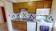 This single-floor unit offers a straightforward, functional layout arranged along a linear floor plan. The kitchen is equipped with warm oak cabinetry — both upper and lower — providing ample storage space. A granite-style laminate countertop runs along the workspace, incorporating a stainless steel sink. The tiled backsplash features a distinctive checkered pattern in white and gray, adding a subtle design element. White appliances include a gas range with overhead microwave, and a dishwasher beneath the counter.

The living area is generously sized, with neutral-toned carpet underfoot and cream-colored walls throughout. A pendant light fixture illuminates the space, and a warm wood-toned entry door serves as a focal point near the foyer, where tile flooring transitions to carpet. The bedroom features soft carpeting and a ceiling fan with light kit, keeping the room comfortable year-round.

The bathroom includes a white vanity with chrome fixtures, a medicine cabinet with mirrored door, and overhead lighting. A hallway connects the rooms, maintaining an organized flow throughout the unit. Windows in the bedroom and living areas allow natural light to enter the space.