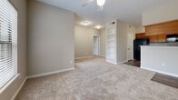 This single-floor residence offers a thoughtfully designed layout with a neutral, warm color palette throughout. The living area is generously sized, featuring plush carpet, ceiling fan, and a built-in shelving unit that adds both storage and character. A pass-through opening connects the living space to the kitchen, which is laid out in a galley configuration with warm wood-toned cabinetry, dark granite-style countertops, wood-look vinyl flooring, and black appliances including a range, microwave, and dishwasher. The bedrooms are carpeted and well-lit, with one room featuring a ceiling fan and a window fitted with horizontal blinds that allow natural light while maintaining privacy. Another bedroom offers a large window with outdoor views. A spacious walk-in closet provides ample storage with wire shelving spanning multiple walls. The bathroom is clean and functional, featuring a full tub and shower combination with white tile surround, wood-look flooring, and overhead cabinetry for additional storage. White six-panel doors and crisp white baseboards complement the neutral taupe walls found throughout, creating a cohesive and move-in-ready living environment.