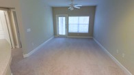 This single-story space features a open living area finished with neutral, light-toned carpeting that extends throughout the room. The walls are painted in a soft beige-green hue, complemented by clean white baseboards that frame the perimeter. A ceiling fan with a light fixture is centrally positioned overhead, providing both airflow and ambient lighting to the space.

Natural light is a notable feature of this room, entering through a large window fitted with horizontal blinds on the far wall. Adjacent to the window is an exterior door with a glass panel, also dressed with blinds, which allows for additional light while offering access to the outdoors. Together, these openings create a bright and airy atmosphere throughout the day.

The layout is straightforward and functional, offering a generous open floor plan that provides flexibility for various furniture arrangements. The space is clean and vacant, making it easy to envision a range of interior configurations. Overall, the room presents a simple, well-maintained environment with practical features suited for comfortable everyday living.