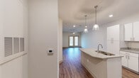 This well-appointed single-floor unit offers a thoughtfully designed open-concept layout that seamlessly connects the kitchen, dining, and living areas. The kitchen serves as a central focal point, showcasing a large granite-topped island with an integrated sink, elegant pendant lighting, crisp white cabinetry, and a full suite of stainless steel appliances including a refrigerator, range, and microwave. Wood-look flooring runs throughout the entire unit, adding warmth and visual continuity across every room. The spacious living area is bathed in natural light courtesy of a set of French doors that open outward, complemented by a ceiling fan overhead. A smart thermostat panel is conveniently mounted near the entryway. The bedroom features a dedicated walk-in closet with built-in shelving, providing generous storage. The bathroom is clean and functional, equipped with a combined tub and shower with chrome fixtures and built-in shelving. A dedicated in-unit laundry room houses a full-size top-loading washer and dryer with overhead wire shelving for additional storage. Recessed lighting throughout enhances the bright, airy ambiance, making this unit a practical and comfortable living space.