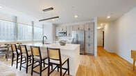 Located at 55 Public Square in the heart of Cleveland, Ohio, this single-floor residence offers a thoughtfully designed, open-concept layout finished with light hardwood flooring throughout the main living areas. The contemporary kitchen is a standout feature, equipped with stainless steel appliances, a large island with seating for multiple guests, white quartz countertops, gray shaker-style cabinetry, a white subway tile backsplash, and a sleek matte black faucet — all illuminated by a modern linear pendant light and large windows that invite natural light. The kitchen flows seamlessly into the living and dining area, where a ceiling fan, recessed lighting, and a cozy fireplace contribute to a comfortable ambiance. A dedicated in-unit laundry area with a full-size washer and dryer adds convenience. The bedroom is carpeted and bathed in natural light through expansive windows that frame captivating views of the Cleveland skyline. A secondary living or flex space also enjoys generous window coverage and city views. Well-lit hallways with recessed lighting and clean white walls connect each room, reinforcing the residence's modern, cohesive aesthetic throughout.
