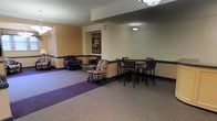 This single-floor lounge space offers a versatile and inviting environment suited for both relaxed socializing and casual dining. The layout is thoughtfully divided into two distinct functional zones. The seating area features a mix of upholstered armchairs and a loveseat with a bold patterned fabric, arranged around a glass-top coffee table, creating an intimate setting for conversation and leisure. A decorative framed vintage-style "Florio" poster serves as a focal point on the wall, adding a touch of character to the space. Adjacent to the lounge area, a dining zone is furnished with tall high-top tables and matching bar-height chairs in a sleek dark finish, ideal for casual meals or informal meetings. The walls are painted in a warm neutral tone with a contrasting dark decorative border running horizontally, contributing to the space's defined aesthetic. Carpeting in complementary tones of purple and beige delineates each zone, adding warmth underfoot. Overhead, flush-mounted and pendant lighting provides consistent, ambient illumination throughout the space. The overall atmosphere is comfortable and functional, reflecting a communal lounge designed to accommodate a variety of activities.