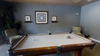 This single-floor space is designed with leisure and entertainment in mind. The standout feature is a generously sized pool table with a light-colored felt surface, complete with a full rack of billiard balls and cues, making it an ideal centerpiece for social gatherings and recreation. The room is finished with cool-toned, slate-blue walls that create a calm and focused atmosphere well-suited for gameplay. Three framed wall pieces are thoughtfully arranged above a pair of floating shelves, adding a decorative touch that enhances the room's character without overwhelming the space. The layout provides ample clearance around the pool table, ensuring comfortable play from all sides. A couch or seating area is partially visible along one wall, offering a spot for onlookers or players awaiting their turn. The overall design balances functionality with a relaxed aesthetic, making this space a well-appointed retreat for recreational activities. The combination of practical layout, tasteful décor, and purpose-driven furnishings makes this an inviting and highly functional game room.