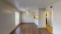 This single-story unit is thoughtfully laid out across several distinct living areas, each finished with consistent, coordinated materials. The main living area greets you at the entry with rich, dark wood-look flooring that extends throughout the common spaces. Natural light filters in through a large window fitted with blinds, while a ceiling fan overhead provides both illumination and airflow. A textured popcorn ceiling runs throughout the unit, adding a characteristic touch to each room.

The kitchen is accessible via a pass-through opening from the living area, offering dark cabinetry contrasted by granite-style countertops and a double basin sink. Black appliances, including a full-size refrigerator and range, complete the functional kitchen setup. Wood-look flooring continues seamlessly into this space.

The bedroom features plush carpeting, a ceiling fan, and a generously sized window that allows ample natural light and an exterior view. The bathroom is well-appointed with a dark-framed vanity mirror, granite-style countertop, dark cabinetry, and a shower-tub combination. The flooring in the bathroom mirrors the wood-look style found in the main living areas, tying the overall design together cohesively.