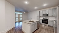 This thoughtfully designed 1-bedroom, 1-bathroom residence offers a clean, modern aesthetic across a single floor. The open-concept layout seamlessly connects the kitchen and living area, creating an airy and functional living space. The kitchen is a standout feature, showcasing gray shaker-style cabinetry, granite countertops, a center island with an undermount sink and built-in dishwasher, and a full suite of stainless steel appliances including a range, microwave, and refrigerator. A subway tile backsplash and wood-look flooring complete the polished look. The living area benefits from abundant natural light streaming through floor-to-ceiling French doors, which also provide access to an outdoor area. A ceiling fan adds comfort to the shared living space. The bedroom is generously sized, finished with plush carpet, a ceiling fan, and a window that brings in additional natural light. It connects directly to a walk-in closet outfitted with wire shelving systems for ample storage. The bathroom features a granite-topped vanity with gray cabinetry, a full tub-shower combination, and wood-look flooring that ties the space together cohesively. Throughout, the residence maintains a bright, neutral palette with white walls and clean finishes.