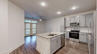 This thoughtfully designed 1-bedroom, 1-bathroom residence offers a clean, modern aesthetic across a single floor. The open-concept layout seamlessly connects the kitchen and living area, creating an airy and functional living space. The kitchen is a standout feature, showcasing gray shaker-style cabinetry, granite countertops, a center island with an undermount sink and built-in dishwasher, and a full suite of stainless steel appliances including a range, microwave, and refrigerator. A subway tile backsplash and wood-look flooring complete the polished look. The living area benefits from abundant natural light streaming through floor-to-ceiling French doors, which also provide access to an outdoor area. A ceiling fan adds comfort to the shared living space. The bedroom is generously sized, finished with plush carpet, a ceiling fan, and a window that brings in additional natural light. It connects directly to a walk-in closet outfitted with wire shelving systems for ample storage. The bathroom features a granite-topped vanity with gray cabinetry, a full tub-shower combination, and wood-look flooring that ties the space together cohesively. Throughout, the residence maintains a bright, neutral palette with white walls and clean finishes.