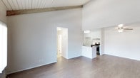 This single-floor space showcases a thoughtfully designed open-concept layout that creates a seamless flow between the living area and kitchen. The most striking architectural feature is the vaulted ceiling with an exposed wooden beam, adding warmth and character to the otherwise clean, modern aesthetic. The soaring ceiling height enhances the sense of spaciousness throughout the main living area.

The flooring throughout is a consistent medium-toned hardwood, providing a cohesive and contemporary look. The walls are finished in a crisp, neutral white, allowing natural light to reflect freely and brighten the interior.

The kitchen is positioned as a natural focal point, featuring a peninsula that doubles as a breakfast bar or workspace. White cabinetry, under-cabinet lighting, and stainless steel appliances contribute to a clean, functional design. A range hood is installed above the stove, combining practicality with a polished finish.

A doorway visible from the main living area leads to additional rooms, suggesting a well-organized floor plan. The overall ambiance is modern and airy, with a minimalist sensibility that makes the space feel open, versatile, and move-in ready.