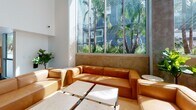 Located in the heart of Los Angeles at 1120 W 6th St, this contemporary amenity lounge is thoughtfully designed to offer both comfort and functionality in a single, well-appointed space. The layout encompasses multiple distinct zones that flow seamlessly into one another, all unified by polished concrete floors and a clean, modern aesthetic.

The centerpiece of the space is a sunlit lounge area featuring soaring floor-to-ceiling windows framed by exposed concrete columns. These expansive glass panels flood the interior with abundant natural light and offer lush views of swaying palm trees outside, creating a bright, resort-like ambiance. Warm cognac leather sofas and sectionals are arranged around minimalist coffee tables, providing inviting seating throughout.

A dedicated recreation zone includes a classic skee-ball lane set against slatted wood panel walls, adding a playful and social dimension to the space. Adjacent to this is a fully equipped kitchen and bar area outfitted with natural wood cabinetry, white quartz countertops, a zellige tile backsplash, brass fixtures, stainless steel appliances — including a large French door refrigerator, dishwasher, and built-in microwave — all illuminated by elegant brass wall sconces. The kitchen is designed for both practicality and style, making it ideal for gatherings and events.