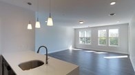 This single-floor space showcases a thoughtfully designed open-concept layout that effortlessly connects the kitchen and living areas into one cohesive environment. The kitchen features a sleek, light-toned countertop with an integrated sink and a matte dark-finish pull-down faucet, complemented by stylish pendant lighting in a modern conical design with dark metal accents. The open flow into the living area is highlighted by smooth, dark hardwood-style flooring that runs throughout, creating a sense of continuity and warmth. Soft, neutral gray walls provide a clean and versatile backdrop that enhances the overall brightness of the space. Recessed and flush-mount ceiling lighting ensures the interior is well-lit at all times. Large double-pane windows in the living area flood the space with natural light, offering pleasant views of lush greenery outside. The clean lines, neutral palette, and modern fixtures combine to create a polished yet inviting atmosphere, making the space both functional and aesthetically refined. The open layout promotes a sense of spaciousness, ideal for both everyday living and entertaining.