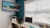 This single-level business center is designed to support productivity in a comfortable, functional setting. The space is anchored by a long, white built-in desk that accommodates two workstations side by side, each equipped with monitors, keyboards, and office supplies. Brown leather-style chairs provide comfortable seating at the shared workspace.

A bold teal accent wall adds a distinctive visual contrast against the light gray walls that define the rest of the space. Warm hardwood flooring runs throughout, contributing to an inviting atmosphere that balances professional utility with aesthetic appeal. White horizontal blinds on the windows allow for adjustable natural light.

The business center also includes multiple printers and a wall-mounted flat-screen television, making it suitable for both individual tasks and monitoring news or media. A small round table with chairs offers an informal meeting or collaborative area within the same room.

A glass-paneled entry door provides views of a landscaped outdoor area and allows additional natural light into the space. Decorative metallic wall art adds a refined finishing touch. The layout is efficient and thoughtfully organized, making this business center a well-rounded resource for professional needs.