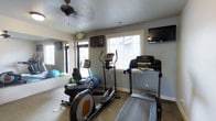 This single-story fitness room offers a practical and functional workout space designed to support a range of cardio-focused exercise routines. The room is equipped with several key pieces of cardio equipment, including a recumbent bike, an elliptical cross-trainer, and two treadmills, providing users with multiple options for low-impact and moderate-intensity workouts. Two wall-mounted flat-screen televisions are positioned at opposite ends of the room, allowing exercisers to stay entertained during their sessions. Natural light filters in through multiple windows fitted with white horizontal blinds, brightening the space and creating an open, airy feel. A ceiling fan overhead aids in air circulation and comfort during use. A large mirror along one wall visually expands the room and is accompanied by a selection of stability and exercise balls in blue and green, adding versatility for stretching or balance training. The textured ceiling and neutral gray walls give the space a clean, no-frills aesthetic. Carpeted flooring runs throughout, providing a degree of cushioning underfoot. The room's layout is efficient, making good use of the available floor space while keeping equipment accessible.