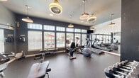 Located at 8001 W Parkway Blvd in Sand Springs, Oklahoma, this fitness center is a well-equipped, single-room exercise facility designed to support a well-rounded workout routine. The space features dark gray walls paired with durable rubber flooring, creating a practical and focused environment for physical activity.

Natural light pours in through a generous bank of large windows fitted with horizontal blinds, offering views of an outdoor pool area complete with seating and colorful umbrellas. Stylish pendant lights and decorative ceiling fans provide additional illumination and complement the modern aesthetic.

The cardio section is equipped with treadmills, elliptical trainers, and a recumbent bicycle, all positioned to take advantage of the poolside views. On the strength training side, the room includes a multi-function cable machine, an adjustable weight bench, and a dumbbell rack stocked with free weights, along with exercise balls for flexibility work. A large wall mirror expands the sense of space and allows for form-checking during workouts.

A wall-mounted flat-screen television adds entertainment for cardio sessions. The room is accessed through a paneled white door with a sidelight window. Exercise room rules are posted near the entrance, and the overall layout is efficiently organized to maximize the usable space within this compact but functional fitness facility.