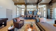 This single-floor club room is a well-appointed communal space designed for relaxation and socializing. The interior showcases soaring ceilings with striking exposed wood beams and a warm wood-paneled ceiling that lend a rustic yet refined character to the space. Wood-look flooring runs throughout, complementing the earthy, neutral color palette and cohesive design aesthetic.

The main lounge area is furnished with plush leather seating, including a sofa and armchairs accented with colorful throw pillows, arranged around a unique live-edge coffee table. A wall-mounted flat-screen television anchors the central gathering zone. Adjacent to the lounge, a bar-height counter with upholstered stools sits beneath pendant lighting, creating an inviting spot for casual dining or conversation.

A dedicated game area features a full-size shuffleboard table accompanied by leather club chairs, offering a relaxed recreational retreat. A large round chandelier adds a sophisticated touch to this section of the room.

Expansive windows dressed with wide-striped drapes flood the space with natural light, while high-top bistro tables positioned near the glass provide views of the outdoor pool area and surrounding community. The overall ambiance is warm, modern, and social — thoughtfully designed for comfortable communal living.