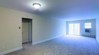 This single-floor residence offers a bright and open layout designed for comfortable everyday living. The main living area is finished with wall-to-wall carpeting in a neutral tone, creating a warm and inviting atmosphere throughout. Smooth, white walls and clean white baseboards contribute to a crisp, well-maintained aesthetic that feels fresh and move-in ready. A flush-mount ceiling light fixture provides consistent overhead illumination, complementing the natural light that pours in through a large sliding glass door along one wall. The sliding door not only brightens the space but also provides convenient access to the outdoors. A doorway leading to an adjacent room or closet is visible, suggesting a functional and connected floor plan that makes efficient use of the available space. The overall ambiance is calm and understated, with a neutral palette that offers a versatile canvas for a variety of interior styles and furnishings. The single-floor layout ensures easy accessibility and a seamless flow between areas, making this a practical and comfortable living environment.