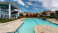 Located at 15000 W Airport Blvd in Sugar Land, Texas, this thoughtfully designed single-floor space combines upscale finishes with an open, welcoming layout. The exterior grounds feature a resort-style swimming pool with a spacious sun deck, a shaded pergola seating area, and lush landscaping, creating an inviting outdoor retreat.

Inside, the expansive living area immediately impresses with its tray ceilings, crown molding, and recessed lighting. Decorative columns define the space while maintaining an open feel. A stone-accented fireplace serves as a focal point, flanked by built-in shelving. Large windows and French doors flood the interior with natural light, and the gleaming large-format tile flooring runs seamlessly throughout.

The gourmet kitchen is a standout feature, centered around an oversized granite-topped island with bar seating for multiple guests. White cabinetry extends to the ceiling, accented by pendant lighting and stainless steel appliances, including a French door refrigerator. A tray ceiling detail adds architectural interest overhead.

The entryway offers a graceful transition into the main living spaces, finished with warm taupe walls, elegant decorative accents, and views directly into the kitchen and living areas. A grand staircase with dark wood railings and a statement chandelier adds further architectural drama to the space.