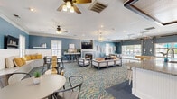 Located at Riverland Woods in Charleston, South Carolina, this generously sized single-floor clubhouse offers a well-appointed, open-concept gathering space designed for comfort and socializing. The interior is defined by a cohesive coastal color palette, with rich slate-blue walls contrasted by crisp white wainscoting and trim throughout.

The main lounge area centers around a classic white-framed fireplace with a mounted flat-screen TV above, flanked by comfortable seating arrangements ideal for relaxed conversation or group viewing. Overhead, ceiling fans provide airflow alongside recessed lighting and a statement chandelier, contributing to a welcoming ambiance.

The full kitchen is a standout feature, showcasing a large granite-topped island with bar seating, stainless steel appliances including a range and refrigerator, white cabinetry, open shelving with warm wood accents, and pendant globe lights overhead. A rustic sliding barn door adds character to the space.

Additional seating zones include built-in window benches with cushions and dining tables paired with industrial-style metal chairs, promoting flexible use of the space. Multiple flat-screen TVs are positioned throughout, and large windows invite natural light and outdoor views. The vibrant geometric-patterned carpet ties the entire space together with a modern coastal flair.