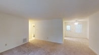 Located at 1201 Columbus Cir in Wilmington, NC, this single-floor home offers a straightforward and functional layout designed for comfortable everyday living. The interior features wall-to-wall carpeting throughout, rendered in warm neutral tones that contribute to a cozy and inviting atmosphere. The main living area is open and airy, divided by a partial interior wall that creates a natural sense of separation between spaces while maintaining an open feel. A ceiling fan is visible in the adjoining area, providing practical climate comfort. Natural light filters in through a windowed entry door, brightening the space and offering a welcoming entrance. The walls are finished in clean white, providing a versatile backdrop that complements a variety of furnishing styles. A hallway visible from the main living space leads to additional rooms, including what appears to be a closet or utility area, suggesting thoughtful storage solutions throughout the home. Recessed floor vents indicate a central HVAC system for year-round comfort. Overall, the home presents a clean, move-in ready environment with a calm, neutral palette well-suited for personalization.