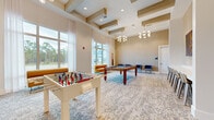 Located at 1799 North Williamson Boulevard in Daytona Beach, Florida, this single-floor game and social lounge is a well-appointed recreational space designed for communal enjoyment. The room makes a strong architectural impression with its soaring ceilings adorned by a distinctive coffered design featuring bold, gold-toned beam accents. Overhead, elegant globe chandeliers with brass fixtures cast a warm, inviting glow throughout the space.

The lounge is centered around entertainment, housing a full-size pool table set atop a patterned area rug, and a foosball table positioned to encourage friendly competition. A wall-mounted flat-screen television is paired with a bar-height counter and backless stools, creating an ideal viewing and socializing zone. Additional seating includes modern navy accent chairs and a sleek mustard-yellow chaise lounge, adding pops of color against the neutral palette.

Floor-to-ceiling windows along one wall flood the interior with natural light while offering open views of the surrounding Florida landscape, complemented by sheer white drapery. One wall features warm wood-toned paneling, lending texture and a refined aesthetic to the overall design. Soft, patterned carpeting runs throughout, tying together the contemporary and comfortable atmosphere of this vibrant social gathering space.
