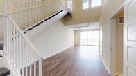 This modern loft-style unit offers a striking open-concept layout defined by soaring double-height ceilings that create an impressive sense of vertical space in the main living area. A prominent staircase with white metal railings ascends to an upper mezzanine level, adding architectural character and visual depth to the interior. Rich, hardwood-style flooring runs throughout, complementing the warm, neutral-toned walls and contributing to a cohesive, contemporary aesthetic.

The living area is bathed in natural light courtesy of expansive floor-to-ceiling windows and a sliding glass door, while track lighting overhead provides additional illumination. The kitchen is thoughtfully designed with rich, dark wood cabinetry extending to the ceiling for ample storage, a light-toned tile backsplash, and a full suite of stainless steel appliances including a refrigerator, electric range, built-in microwave, and dishwasher. Light stone countertops and a single-basin sink complete the functional workspace.

The bathroom features a dark wood vanity with a wide mirror and integrated lighting, a full tub-and-shower combination with white tile surround, and hardwood-style flooring that ties back to the rest of the unit. Additional rooms provide versatile, open spaces illuminated by track lighting, offering flexibility for various uses.