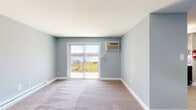 This single-floor unit is characterized by a consistent soft blue-gray color palette throughout, creating a calm and cohesive atmosphere across all rooms. The carpeted living area is generously proportioned, featuring a sliding glass door that opens to a private balcony overlooking what appears to be a scenic waterfront setting, allowing natural light to fill the space. A wall-mounted air conditioning unit and baseboard heating provide year-round climate control. The layout, as revealed in the overhead floor plan view, includes a living area, kitchen, bedroom spaces, and a bathroom arranged in a practical and accessible configuration.

The kitchen is a functional standout, featuring rich dark wood cabinetry, a mosaic tile backsplash, stainless steel countertops, a double sink, and black appliances including an electric range — all complemented by a window with a water view. The bedrooms are carpeted and equipped with ceiling fans and baseboard heating; one includes a closet and access to adjoining rooms. The bathroom offers a full tub-shower combination with white tile surround and blue-gray floor tile. Throughout the unit, white trim, clean lines, and neutral finishes contribute to a simple, well-maintained aesthetic.