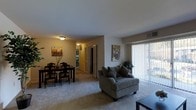 This single-floor residence showcases an open-concept layout that seamlessly connects the living and dining areas, creating a cohesive and functional living space. The living area is anchored by plush wall-to-wall carpeting in a neutral tone, lending warmth and comfort underfoot throughout the space. A large sliding glass door fitted with vertical blinds allows an abundance of natural light to fill the room while providing direct access to an outdoor area.

The dining area is positioned adjacent to the living space and accommodates a full dining table with seating for several guests. Artwork on the walls adds a decorative touch to the otherwise neutral color palette of soft whites and beiges. Ceiling-mounted light fixtures provide ambient illumination throughout both areas, supplementing the natural light from the sliding door.

A hallway visible in the background suggests additional private spaces beyond the main living area, including what appears to be a corridor leading to further rooms. The open flow between the dining and living zones makes the space feel generously proportioned and well-suited for both everyday living and casual entertaining. Overall, the unit presents a comfortable, understated aesthetic with practical layout considerations.