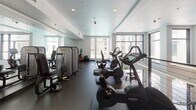 This two-story fitness center is designed with a contemporary aesthetic, characterized by a crisp white interior, dark rubberized flooring, and a dramatic double-height volume on the lower level. Floor-to-ceiling windows and glass entry doors flood the main floor with natural light, offering views of the surrounding outdoor areas and creating an open, energizing atmosphere.

The ground level serves as the primary strength-training zone, housing a multi-station cable machine, adjustable weight benches, a power tower, and a well-stocked dumbbell rack. The soaring ceiling height gives this area a spacious, airy feel, with linear LED lighting accenting the upper walls. A staircase with a sleek metal cable railing connects to the upper level, which features an open mezzanine overlooking the main floor below.

The upper level is dedicated to cardiovascular and machine-based training. It is equipped with treadmills, stationary bikes, ellipticals, and a range of selectorized strength machines. Full-length mirrors on one wall visually expand the space, while recessed lighting and windows maintain a bright, comfortable environment. Additional rooms on the upper floor provide supplementary workout or utility space. Together, both levels offer a well-rounded and professionally equipped facility.