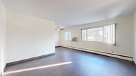 This single-floor residential unit offers a clean, functional layout designed for comfortable everyday living. The expansive main living area is bright and open, featuring rich dark hardwood flooring, crisp white walls, and a ceiling fan overhead. Multiple windows along one wall flood the space with natural light and provide views of the surrounding exterior, while a baseboard heating system and a wall-mounted air conditioning unit ensure year-round comfort.

The galley-style kitchen is efficiently arranged, offering ample white cabinetry, generous counter space with a double sink, and dark hardwood flooring that ties the space together. A full-size refrigerator anchors the kitchen, making it well-equipped for daily use.

The bedroom is a comfortable, carpeted retreat featuring a large window with vertical blinds that allow for adjustable natural light and a baseboard heater for warmth. A wide sliding-door closet provides substantial storage with wood-toned shelving and hanging rods.

The full bathroom is appointed with classic small-format mosaic tile flooring, a pedestal sink, a toilet, and a shower-tub combination enclosed by a curtain. Warm wood-trimmed door framing adds a touch of character. Overall, the space presents a straightforward, well-maintained interior suited for practical, comfortable living.