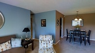 Located at Regency, this single-floor residence offers a well-organized and functional living space. The main living area is characterized by a two-tone color scheme, combining cool slate blue and warm taupe accent walls that create a visually dynamic yet cohesive atmosphere. Carpeted flooring runs throughout, adding warmth and comfort underfoot. The living area is open and accommodating, providing ample room for seating arrangements, as demonstrated by the patterned lounge chair and chaise that serve as a focal point. A dark wood side table with a classic black table lamp adds a grounded, traditional touch to the space. Wall décor, including a framed piece and a circular metal accent, enhances the room's personality. A hallway visible from the living area connects to additional rooms, suggesting a practical and navigable layout. The dining area, visible to the right, features a dark wood table and chairs set beneath a textured ceiling, positioned near a window that introduces natural light. Overall, the space presents a cozy and well-appointed interior with a thoughtful blend of colors and furnishings.