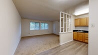Located at 5417 Morris Ave in Suitland, MD, this single-floor, one-bedroom, one-bathroom unit offers a well-organized and functional layout. The living room is generously sized, featuring neutral-toned carpet, cream-colored walls, and a wide set of windows that allow abundant natural light and views of surrounding greenery. A distinctive open-shelving room divider separates the living area from the kitchen, adding a structural design element while maintaining an open feel. The kitchen features warm wood-toned cabinetry and a dark countertop, transitioning to light hardwood-style flooring that complements the overall neutral palette. The bedroom is a comfortable, carpeted space with a wide double window overlooking tree-lined outdoor views and a built-in closet with bifold doors and a hanging rod for organized storage. The bathroom is clean and efficiently designed, with white tile flooring, a full tub-shower combination, a wood-cabinet vanity, and bright overhead lighting. Throughout the unit, the consistent use of neutral tones, carpet in living and sleeping areas, and ample window placement create a cohesive and well-lit living environment suitable for everyday comfort.