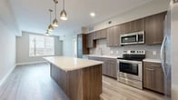 This well-appointed one-bedroom, one-bathroom unit offers a thoughtfully designed open-concept layout with light wood-style flooring that flows seamlessly throughout the space. The combined kitchen and living area forms the heart of the unit, showcasing a contemporary kitchen with flat-panel walnut-toned cabinetry, a geometric tile backsplash, white quartz countertops, and a full suite of stainless steel appliances including a range, built-in microwave, dishwasher, and a large side-by-side refrigerator. A central kitchen island with a white quartz top provides additional prep space and serves as a natural focal point, accented by stylish gold pendant lighting overhead. The adjacent living area benefits from large windows fitted with horizontal blinds, allowing ample natural light and outdoor views, and is complemented by a ceiling fan. The bedroom is a comfortable, well-proportioned room with its own large window overlooking the outdoors, a ceiling fan, and access to a dedicated closet. The bathroom features large-format tile flooring, an extended double-sink vanity with warm wood cabinetry, white quartz countertops, a wide mirror with bar lighting, and a full tub-shower combination with white subway tile surround.