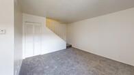 Located at 3541 Evans Ave in Fort Myers, Florida, this two-bedroom, one-bathroom unit is thoughtfully laid out across two floors. The main level houses a combined living room and kitchen area, where the open layout connects the carpeted living space to the kitchen. The kitchen features warm wood-toned cabinetry, white appliances including a refrigerator and electric range, and tile flooring that provides a practical contrast to the carpeted areas. A ceiling fan in the living room adds comfort, and a large window with vertical blinds allows natural light to brighten the space. A closet and entry door are conveniently positioned on the main floor as well. A staircase with a white railing leads to the upper level, which hosts both bedrooms. The primary bedroom offers generous proportions with plush carpet, a wide louvered bi-fold closet, and access to adjoining spaces. The second bedroom is cozy and well-lit by a window with vertical blinds. The bathroom features a vanity with a yellow-toned countertop, a large mirror, and a lighted fixture overhead. Throughout the unit, neutral white walls, textured ceilings, and consistent carpeting create a clean, uniform aesthetic suited for comfortable everyday living.