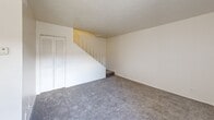 Located at 3541 Evans Ave in Fort Myers, Florida, this two-bedroom, one-bathroom unit is thoughtfully laid out across two floors. The main level houses a combined living room and kitchen area, where the open layout connects the carpeted living space to the kitchen. The kitchen features warm wood-toned cabinetry, white appliances including a refrigerator and electric range, and tile flooring that provides a practical contrast to the carpeted areas. A ceiling fan in the living room adds comfort, and a large window with vertical blinds allows natural light to brighten the space. A closet and entry door are conveniently positioned on the main floor as well. A staircase with a white railing leads to the upper level, which hosts both bedrooms. The primary bedroom offers generous proportions with plush carpet, a wide louvered bi-fold closet, and access to adjoining spaces. The second bedroom is cozy and well-lit by a window with vertical blinds. The bathroom features a vanity with a yellow-toned countertop, a large mirror, and a lighted fixture overhead. Throughout the unit, neutral white walls, textured ceilings, and consistent carpeting create a clean, uniform aesthetic suited for comfortable everyday living.
