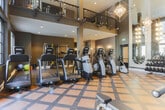This fitness center in Sandy Springs, Georgia, is a sophisticated and comprehensively equipped facility designed to accommodate a wide range of workout preferences. The space makes an immediate architectural impression with its dramatically soaring double-height ceilings and expansive floor-to-ceiling windows framed in dark steel, allowing abundant natural light to pour in while offering views of an outdoor pool and courtyard beyond. Geometric chandelier fixtures and elegant multi-arm gold pendant lights add a refined, upscale ambiance uncommon in typical fitness spaces.

The main floor features a patterned, multi-toned rubber flooring in warm tan and brown tones, providing both visual interest and functional durability. A row of modern elliptical and cardio machines lines the windowed wall, oriented to take full advantage of the outdoor views. The strength-training area is anchored by a full dumbbell rack set against arched floor-to-ceiling mirrors, which amplify the sense of space. Additional equipment includes cable machines, weight benches, and a stationary bike.

A secondary area with darker flooring houses functional training equipment, including a cable resistance tower, medicine ball rack, stability balls, and TRX-style suspension systems, supporting diverse training modalities. Recessed lighting and wall sconces throughout ensure consistent, well-distributed illumination across the entire facility.