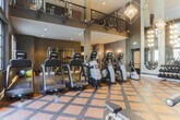 This fitness center in Sandy Springs, Georgia, is a sophisticated and comprehensively equipped facility designed to accommodate a wide range of workout preferences. The space makes an immediate architectural impression with its dramatically soaring double-height ceilings and expansive floor-to-ceiling windows framed in dark steel, allowing abundant natural light to pour in while offering views of an outdoor pool and courtyard beyond. Geometric chandelier fixtures and elegant multi-arm gold pendant lights add a refined, upscale ambiance uncommon in typical fitness spaces.

The main floor features a patterned, multi-toned rubber flooring in warm tan and brown tones, providing both visual interest and functional durability. A row of modern elliptical and cardio machines lines the windowed wall, oriented to take full advantage of the outdoor views. The strength-training area is anchored by a full dumbbell rack set against arched floor-to-ceiling mirrors, which amplify the sense of space. Additional equipment includes cable machines, weight benches, and a stationary bike.

A secondary area with darker flooring houses functional training equipment, including a cable resistance tower, medicine ball rack, stability balls, and TRX-style suspension systems, supporting diverse training modalities. Recessed lighting and wall sconces throughout ensure consistent, well-distributed illumination across the entire facility.