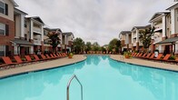 Located at 410 Starmount Park Blvd in Kannapolis, North Carolina, this single-story community clubhouse offers an impressive array of amenities designed for relaxation and socializing. The centerpiece of the outdoor area is a expansive resort-style swimming pool, flanked by generous poolside decking lined with lounge chairs, creating an inviting retreat surrounded by lush landscaping and the community's attractive residential buildings.

Inside, the clubhouse is finished with warm wood-look flooring throughout, lending a cohesive and upscale feel to every area. The lounge and dining space is a standout, anchored by a dramatic stacked-stone accent wall and a bold wooden ceiling beam from which three sculptural metallic globe pendant lights hang, casting a warm, ambient glow over a large dining table with bench and chair seating. Comfortable upholstered seating nearby creates a relaxed gathering atmosphere.

The game room features a full-size billiards table positioned beneath soaring ceilings and an abundance of oversized multi-pane windows that flood the space with natural light and offer direct views of the pool area. An adjoining kitchen and café-style seating area provides convenient space for residents to gather. The overall ambiance is modern yet welcoming, balancing stylish design with practical, community-centered functionality.