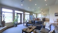 This single-story space is designed with an open-concept layout that seamlessly connects the living, dining, and kitchen areas into one cohesive environment. The living area is furnished with a substantial dark leather sectional sofa and coordinating armchair, arranged around a stone-topped coffee table that anchors the space. Plush carpet underfoot adds warmth throughout the main seating area.

Natural light flows generously through large windows and glass-paneled entry doors, complemented by recessed ceiling lights and a ceiling fan that ensure consistent illumination and airflow. Potted tropical plants near the entry add a touch of greenery and life to the space.

The kitchen, visible from the living area, features warm wood cabinetry, stainless steel appliances including a refrigerator and wall oven, and ample counter space. A breakfast bar with seating connects the kitchen to the dining area, making it ideal for casual gatherings. Neutral wall tones and consistent flooring transitions tie the overall aesthetic together, creating a functional yet inviting atmosphere suited for community or shared use.