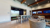 This single-floor entertainment lounge is a thoughtfully designed recreational space that blends modern aesthetics with a wide variety of leisure amenities. The interior is defined by soaring ceilings accented with exposed wood beams and a dark upper ceiling treatment, creating a dramatic yet welcoming atmosphere. Polished concrete floors run throughout, lending a sleek, contemporary feel.

At the heart of the space is a dedicated indoor golf simulator bay, enclosed by dark curtains and equipped with a full hitting mat, large projection screen, and a padded bench seating area for spectators. Adjacent to the simulator, a tall bar-height table with upholstered counter stools serves as a social hub for gathering and viewing.

The main lounge area features a warm wood-paneled accent wall mounted with dual flat-screen televisions, alongside a shuffleboard table for casual gameplay. Comfortable lounge seating—including woven armchairs and plush sofas—is arranged around a wood-stump side table, encouraging relaxed social interaction.

A separate nook offers a classic arcade game cabinet paired with additional seating and decorative shelving with warm wood wall paneling. Large floor-to-ceiling windows and glass doors along one wall flood the space with natural light and provide direct views of an outdoor courtyard. Recessed lighting throughout ensures the space is well-lit and inviting at all hours.