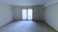 This single-floor space offers a clean, functional layout designed for comfortable everyday living. The main living area is expansive and open, finished with plush wall-to-wall carpeting that adds warmth and a sense of coziness underfoot. Neutral white walls keep the space feeling bright and airy, providing a versatile backdrop that easily accommodates a variety of furnishing styles and décor preferences. A central feature of the living area is a set of sliding glass doors framed by vertical blinds, which serve as both a practical entryway to the outdoors and a generous source of natural light. The doors offer a view of a greenery-lined outdoor area, bringing a touch of nature into the interior. Overhead, a ceiling-mounted HVAC vent ensures year-round climate comfort throughout the space. The overall ambiance is understated and move-in ready, with a simple, well-maintained aesthetic that appeals to those seeking a clean and adaptable living environment. The single-floor layout ensures ease of navigation, making the space accessible and straightforward in its design.