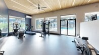 This well-appointed fitness center in Portland, Oregon offers a comprehensive workout environment designed for both functionality and comfort. The space is defined by a standout natural wood-planked ceiling with integrated linear LED lighting, lending warmth and a modern aesthetic throughout. Walls are finished in a two-tone palette of cool navy blue and neutral gray, creating an energizing yet focused atmosphere.

The facility is equipped with a diverse range of cardio and strength-training equipment, including treadmills, stationary and spin bikes, a rowing machine, multi-station cable machines, Nautilus strength machines, and a well-stocked free weights area featuring a full dumbbell rack and kettlebells. A medicine ball and resistance band storage rack adds to the variety of functional training options available.

Durable rubber flooring spans the entire space, providing safety and comfort underfoot. Large windows along multiple walls flood the interior with natural light and offer direct views of the outdoor pool area, creating a bright and motivating environment. A glass door provides convenient access to the pool deck. A ceiling fan and wall-mounted air conditioning unit ensure climate comfort year-round. A large mirror enhances the sense of openness. The fitness center connects to adjacent community amenity spaces, making it a central hub for residents.