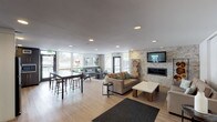 This single-floor community amenity space in Thornton, Colorado offers a well-appointed gathering area designed for comfort and functionality. The open-concept layout features light hardwood flooring throughout, recessed lighting, and large windows that flood the space with natural light while framing views of the surrounding outdoor areas.

The main lounge area is anchored by a striking stacked-stone accent wall housing a sleek linear fireplace and wall-mounted flat-screen TV, flanked by decorative wood-slice art displays. Multiple seating arrangements with neutral-toned sofas and accent pillows create inviting conversation areas, while a high-top bar table with stools provides an additional casual dining or workspace option.

A secondary seating nook near the exterior door features a charcoal sofa set against a textured wallpapered wall, offering a quieter retreat with direct outdoor access and ample window views.

The kitchenette is outfitted with white shaker-style cabinetry, stone countertops, a reflective glass tile backsplash, stainless steel refrigerator, and a deep-set sink — ideal for hosting or everyday use. Adjacent to the kitchenette, a large window overlooks a fully equipped fitness center, connecting the social and wellness amenities of the building seamlessly.
