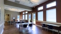 This single-story space offers a versatile, open interior designed to accommodate a variety of functions, from collaborative work sessions to classroom-style gatherings. The room is defined by its notably high ceilings, which create an expansive, airy atmosphere and contribute to a sense of openness throughout. Rich brown accent walls contrast with lighter gray tones and white trim, lending the space a modern yet welcoming aesthetic.

Natural light is a standout feature, with multiple large windows positioned both at standard height and as upper transom windows near the ceiling line, flooding the interior with daylight and offering views of the surrounding greenery. Several glass-paneled doors provide direct access to the outdoors, reinforcing the connection to the exterior landscape.

The polished concrete floor anchors the space with a clean, durable finish, while the drop ceiling with recessed ventilation and flush lighting ensures practical functionality. Furnishings include lightweight rectangular tables and black stackable chairs arranged in a flexible configuration, allowing easy reconfiguration to suit different uses. A long counter along one wall provides additional workspace. The overall ambiance is bright, organized, and well-suited for productive, community-oriented activities.