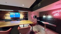 This single-floor clubhouse is designed as a vibrant, multifunctional social space with a carefully curated aesthetic that blends modern industrial elements with playful, lounge-inspired décor. The interior is anchored by an exposed ceiling with dark ductwork and geometric copper-toned framework overhead, lending an industrial-chic character to the space.

At the heart of the clubhouse is a generously sized bar area featuring a live-edge wood communal table surrounded by stylish rust-toned swivel chairs. A large flat-screen television is mounted against a warm wood-paneled accent wall, complemented by a sleek dark countertop with built-in beverage refrigeration below — ideal for entertaining and social gatherings.

Adjacent to the bar, a lounge zone features tufted black vinyl booth seating set against a pink marble-style accent wall. A standout neon sign reading "pump up the volume" adds a bold, energetic focal point. Arc floor lamps and ambient pink lighting enhance the moody, nightlife-inspired atmosphere.

The space extends into a game room area equipped with a classic shuffleboard table and a large wall-mounted Scrabble board. A pair of blue armchairs offers additional casual seating nearby. Tile flooring runs throughout, tying the entire clubhouse together cohesively.