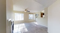 This single-floor space offers an open and airy layout that maximizes both comfort and functionality. The expansive living area is finished with soft carpeting and features a ceiling fan with a light fixture, providing year-round comfort and warmth. Large windows along the exterior wall flood the room with generous natural light, casting a bright and welcoming ambiance throughout the space.

The living area flows seamlessly into a dining and kitchen zone, connected through an open pass-through counter that enhances the open-concept feel. The kitchen is equipped with dark wood cabinetry, a refrigerator, and overhead lighting, offering a practical and efficient workspace. A pendant light above the counter adds a subtle decorative touch to the transition between spaces.

The textured popcorn ceiling adds a classic character to the interior, while the neutral beige wall tones create a versatile backdrop suitable for a variety of décor styles. Tile flooring in the kitchen and dining area contrasts with the plush carpeting in the living space, defining each zone distinctly. Overall, the layout is thoughtfully designed to promote an easy flow between areas, making this a bright, functional, and inviting single-floor living space.